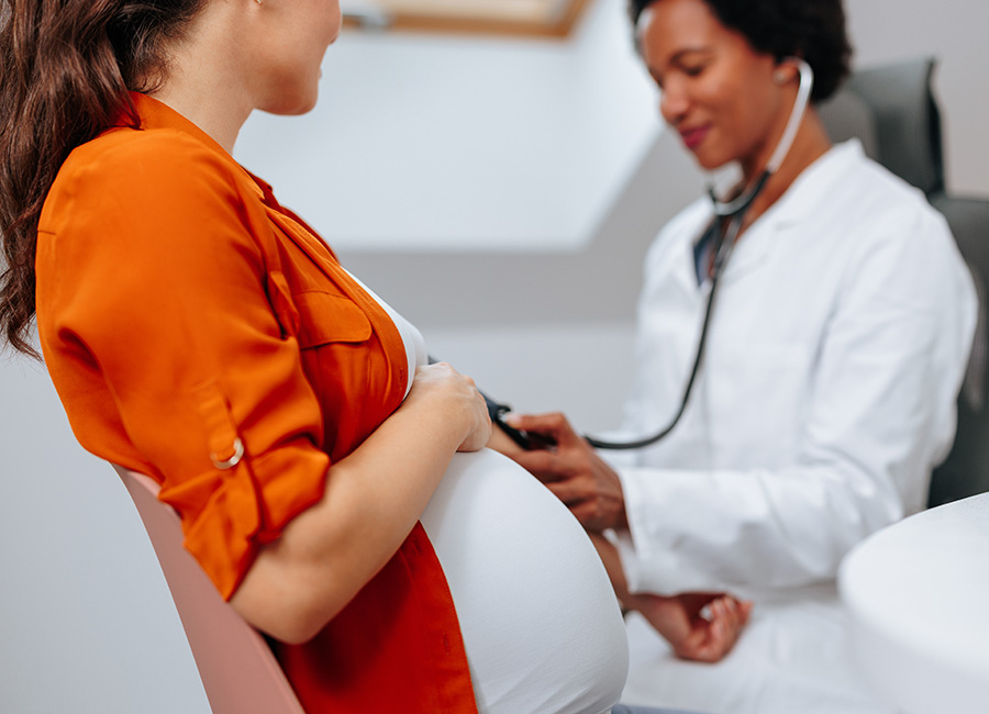 A female doctor listens to the belly of a pregnant woman wearing a bright orange shirt.