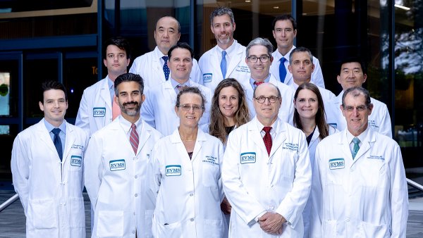 A group of ear, nose, and throat doctors in white coats stand in front of a glass office building named Waitzer Hall.